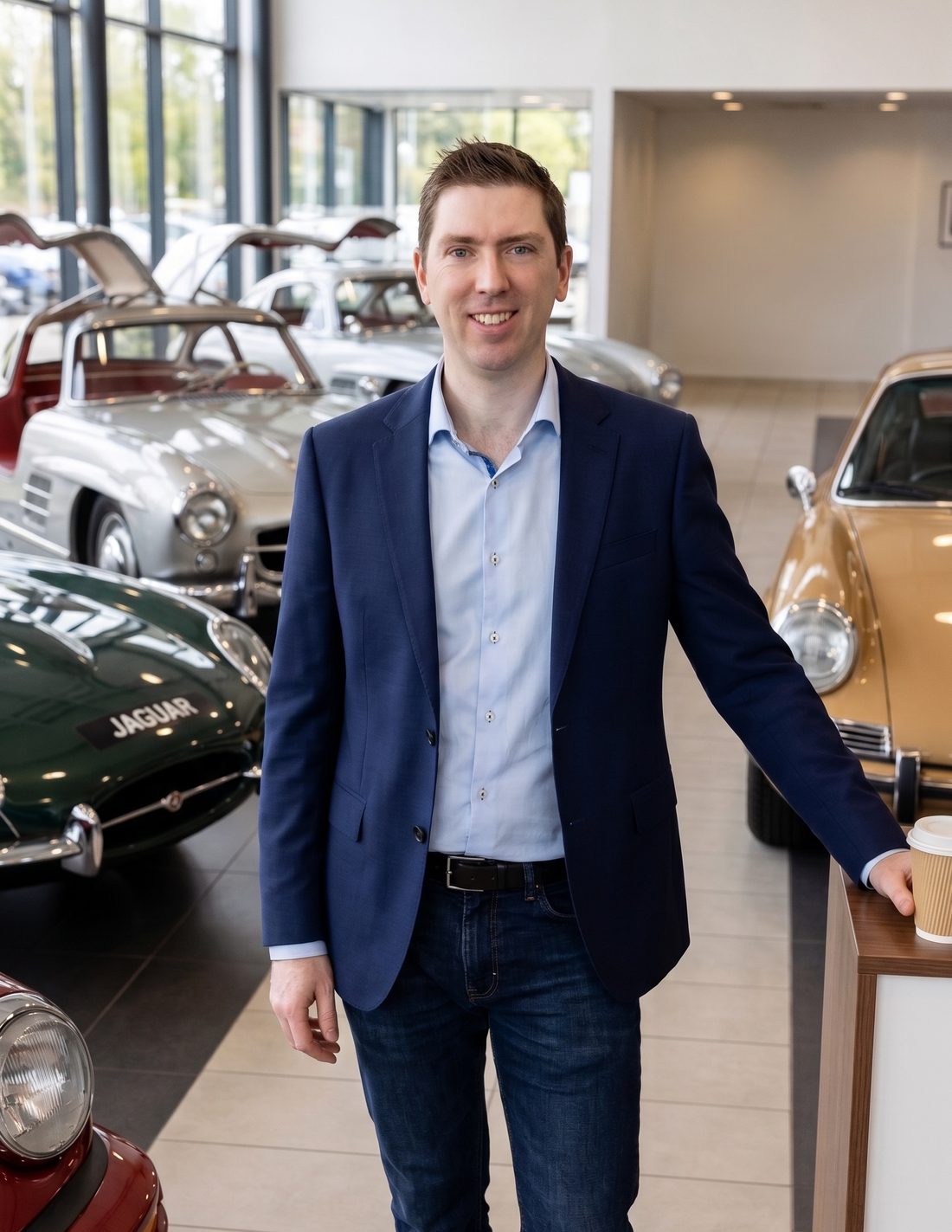 Glenn Harwood in a classic car showroom with Mercedes 300SL Gullwings, a Jaguar E-Type, and a Porsche 911